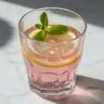 Pink salt recipe drink in clear glass with lemon wheel and mint garnish on white marble surface
