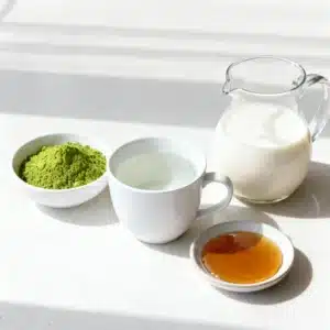 Modern flat lay photo of the ingredients for the perfect matcha tea recipe: vibrant green matcha powder in a small white bowl, white ceramic cup with hot water, glass jug of oat milk, and small dish of maple syrup on a light-colored countertop.