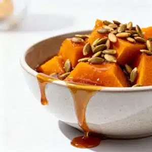 A serving of warm Calabaza en Tacha in a white bowl, drizzled with thick syrup and garnished with crunchy toasted pepitas.