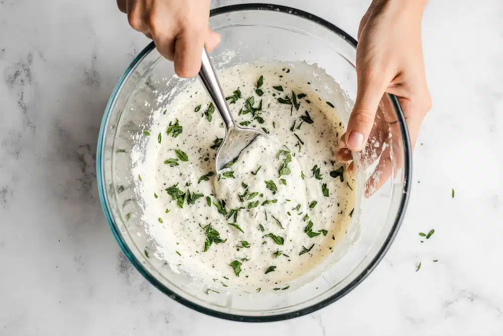 Hands mixing boursin cheese and herbs in a modern glass bowl
