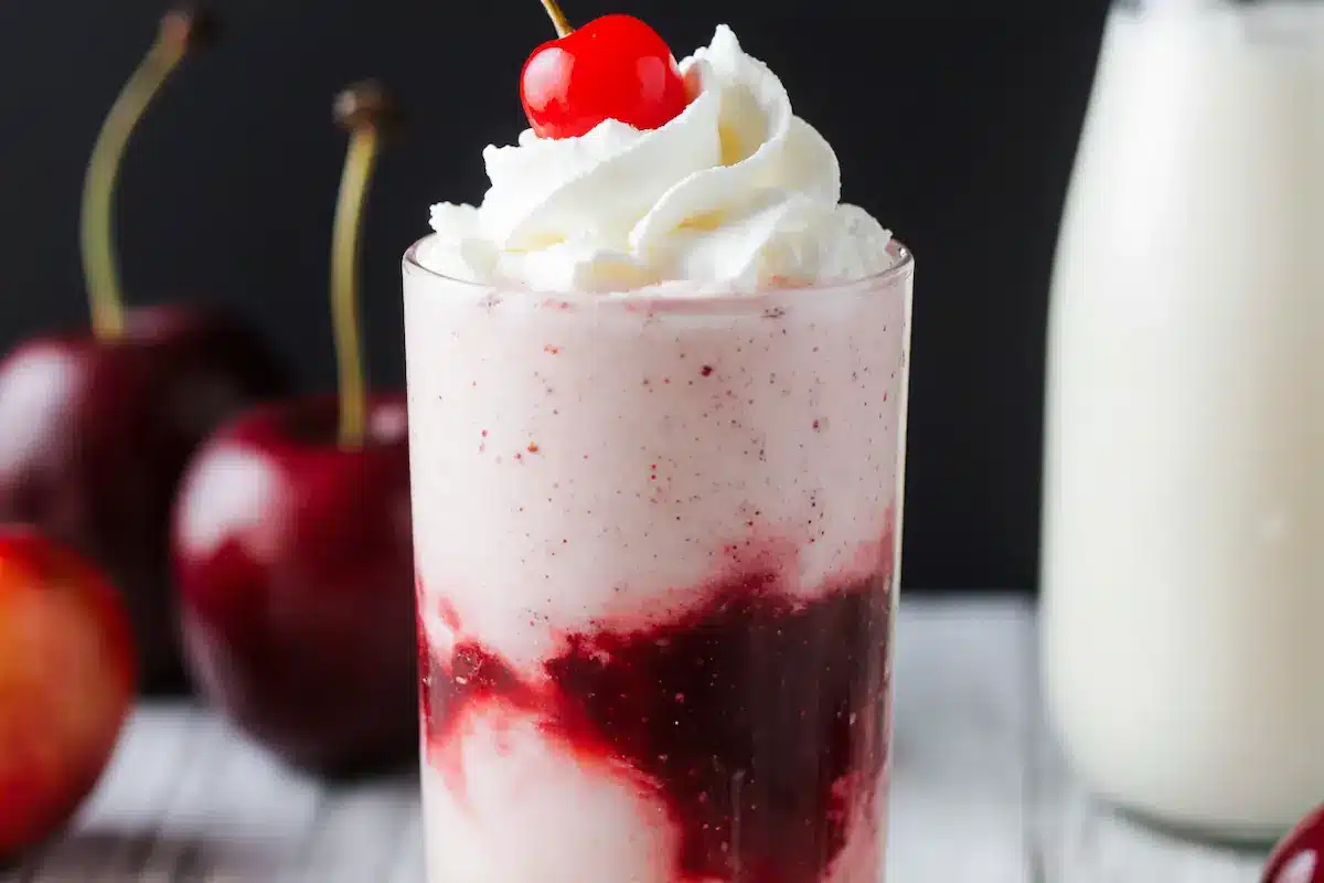 A tall glass of homemade cherry cream soda, perfectly layered and garnished with whipped cream and a cherry.